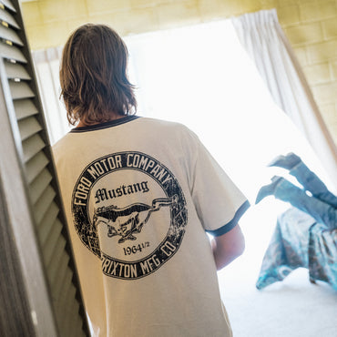 A person viewed from behind wearing a cream-colored ringer t-shirt with a large black circular Ford Mustang graphic on the back, standing in a brightly lit room.