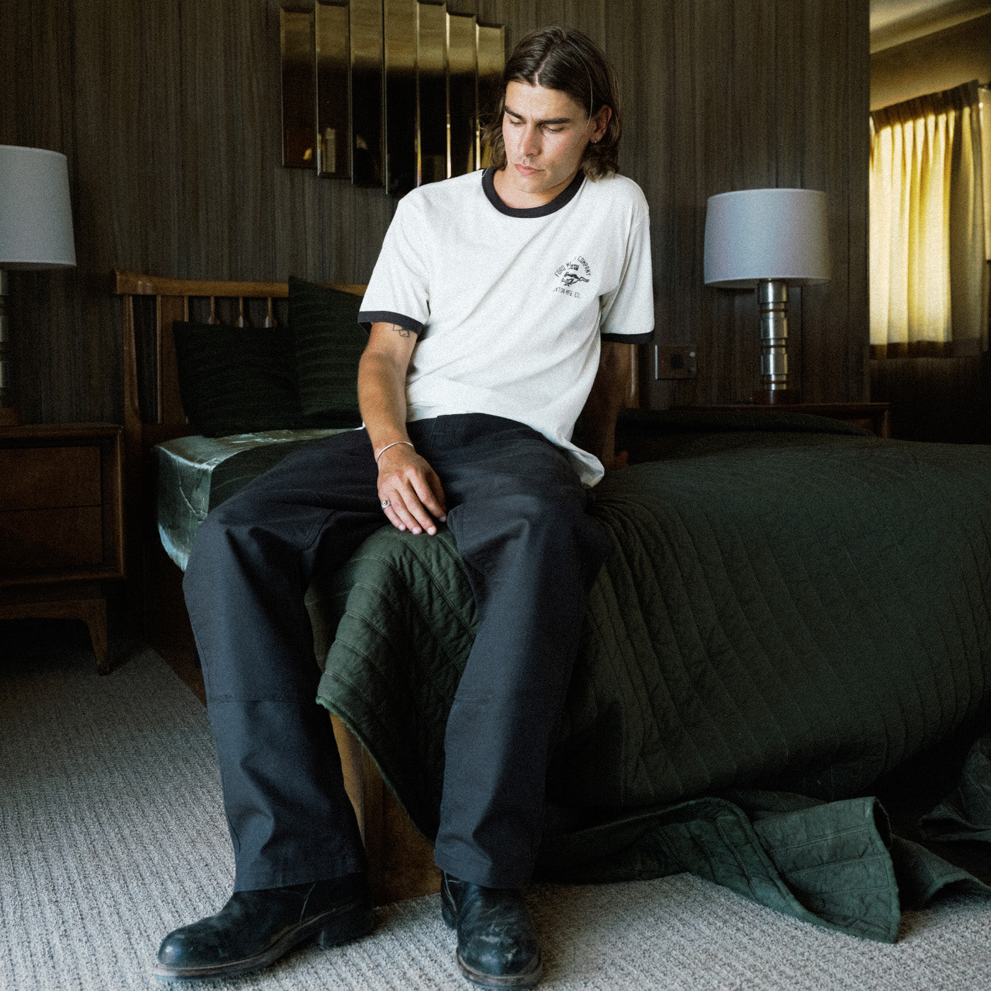 A man sits on a bed in a rustic bedroom setting, wearing a white crewneck t-shirt with black trim and a small graphic on the left chest, paired with dark charcoal pants and black boots.
