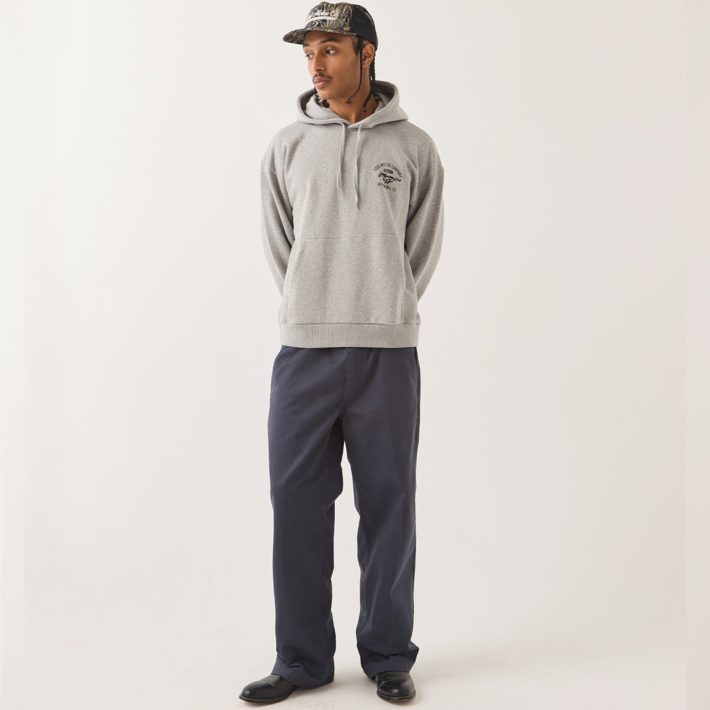 A man wearing a light gray heather pullover hoodie with a small black chest graphic, dark navy pants, and a camouflage baseball cap, standing against a plain off-white studio background.