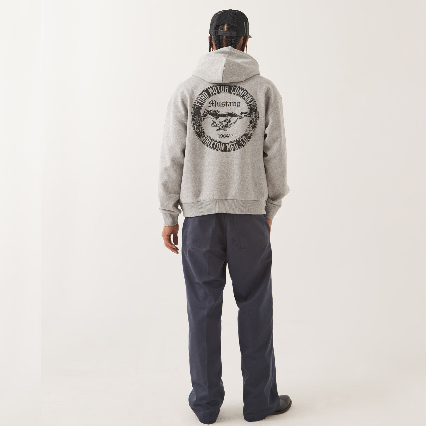 A man viewed from behind wearing a light gray heather pullover hoodie with a large black circular graphic on the back, paired with dark navy pants and a black cap.