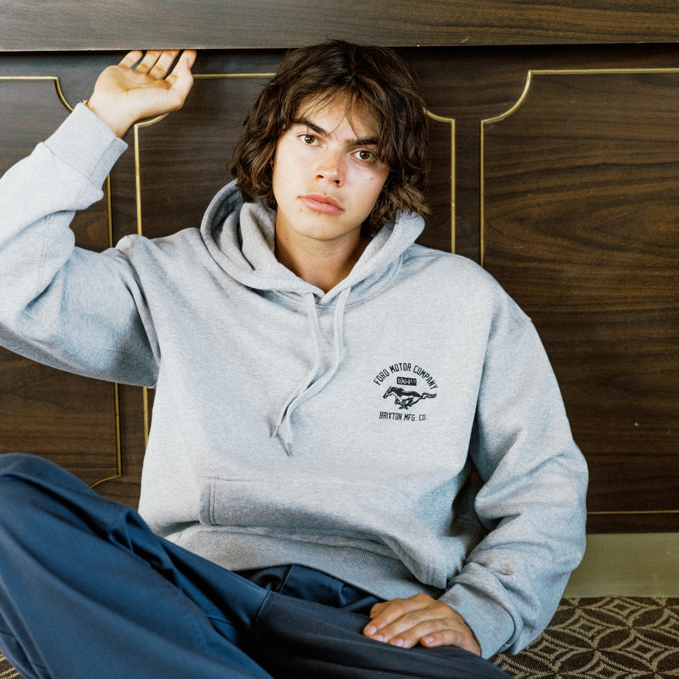 A person wearing a light gray pullover hoodie with a black graphic on the left chest, seated against a dark wood-paneled background.