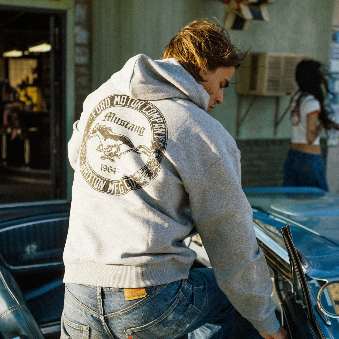 A person wearing a light gray hoodie with a large vintage-style Ford Mustang back graphic, leaning over a blue car in an outdoor setting.