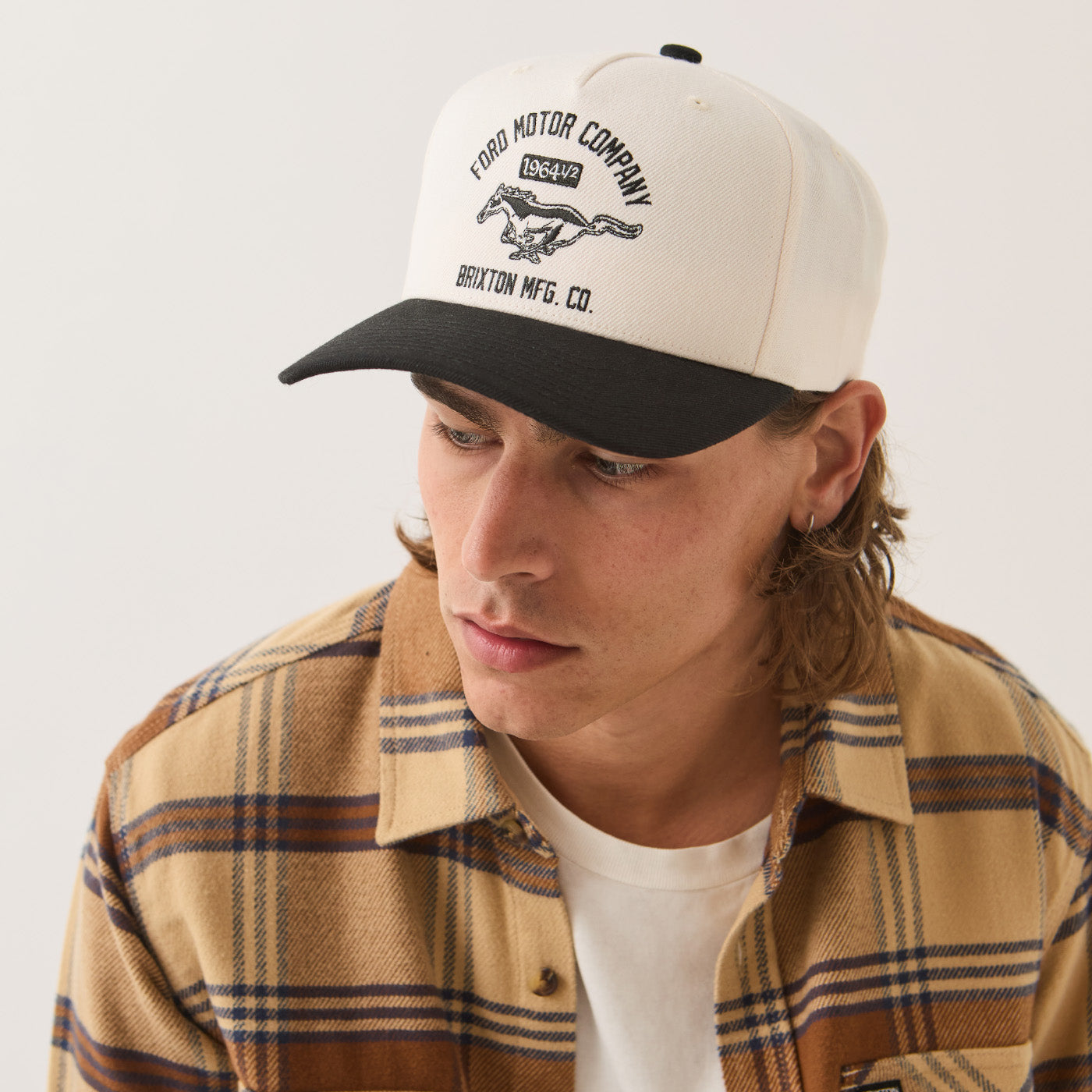 A man wearing a Ford Motor Company 1967 Mustang baseball cap with a cream crown and black brim, paired with a tan and brown plaid flannel shirt over a white t-shirt, photographed against a light off-white background.