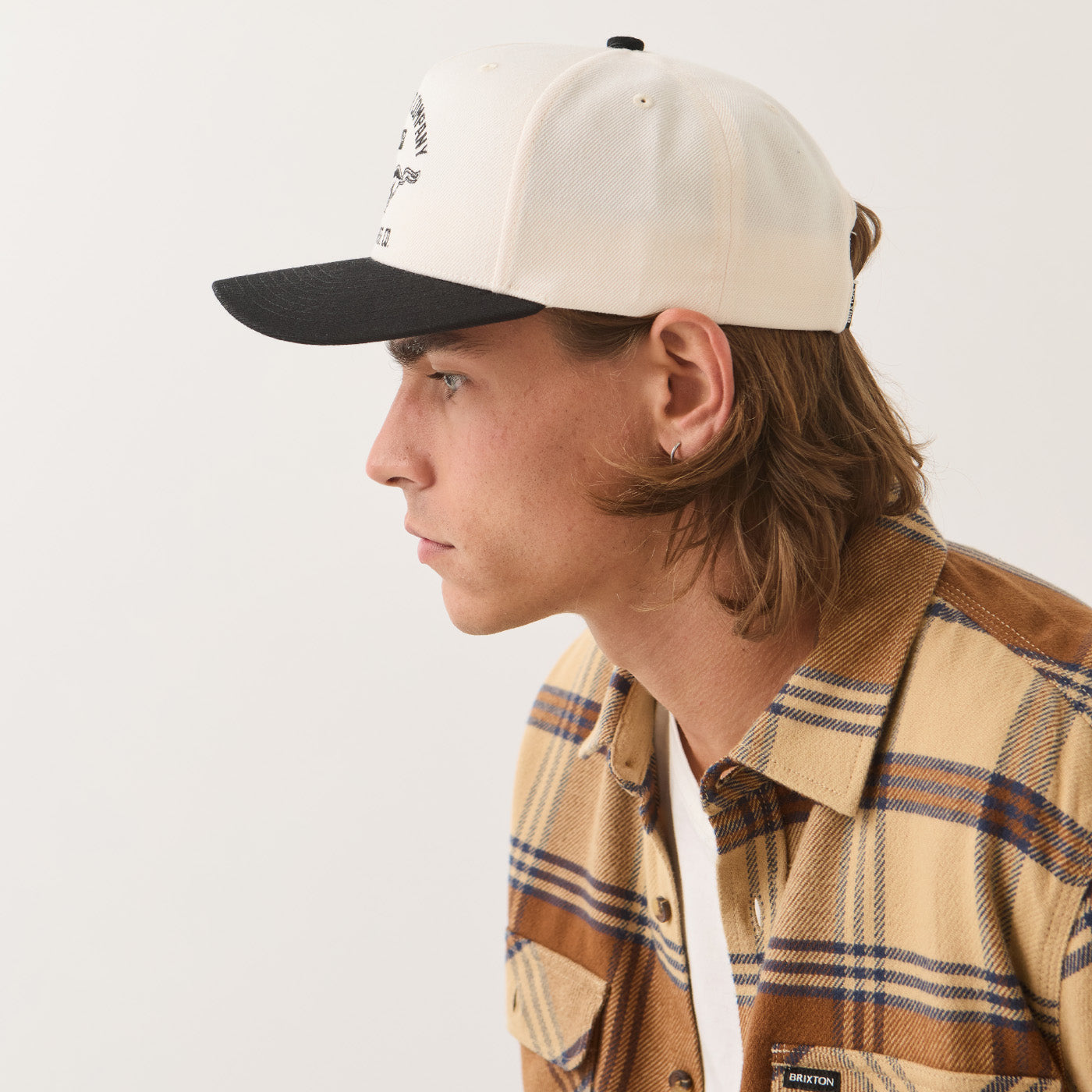 A side-profile portrait of a young man wearing a cream and black baseball cap with a tan and brown plaid button-up shirt against a plain off-white studio background.