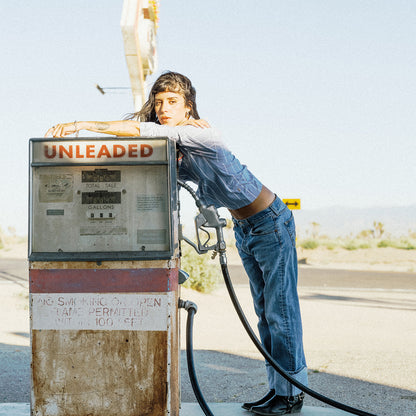 A woman leans against a vintage gas pump at an outdoor gas station, wearing a blue denim shirt and jeans. The pump has a prominent 'UNLEADED' sign on top.