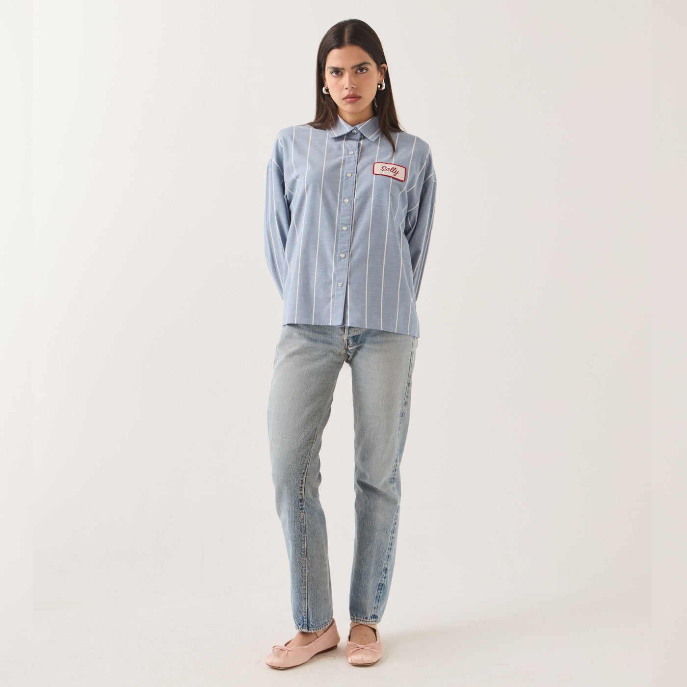 A model stands against a plain light background wearing a light blue and white vertically striped button-up shirt with a pink embroidered patch on the chest, paired with light-wash jeans and pale pink flats.
