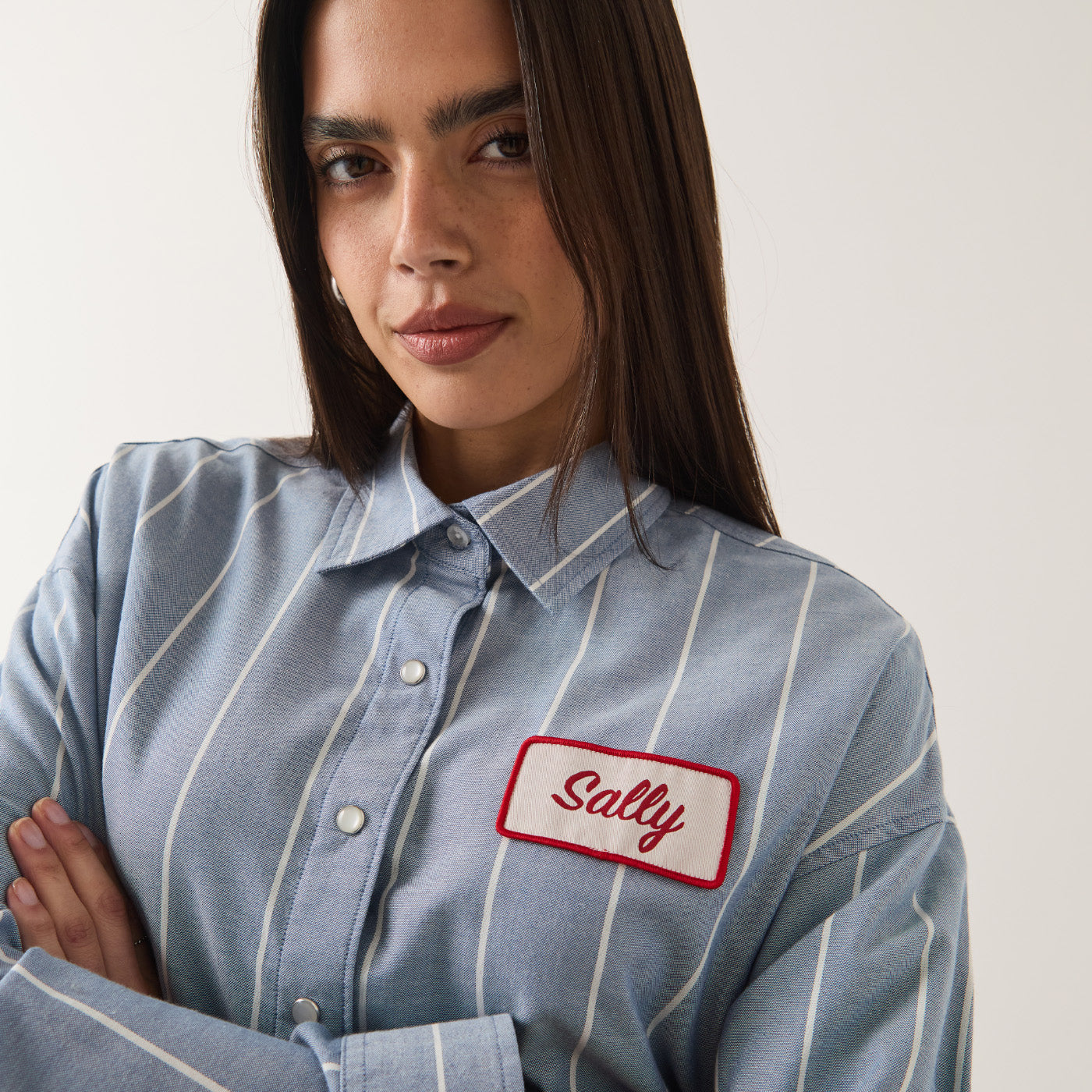 A woman wearing a light blue and white vertically striped button-up shirt with a white rectangular name patch on the chest that reads 'Sally' in red script with a red border.
