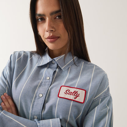 A woman wearing a light blue and white vertically striped button-up shirt with a white rectangular name patch on the chest that reads 'Sally' in red script with a red border.