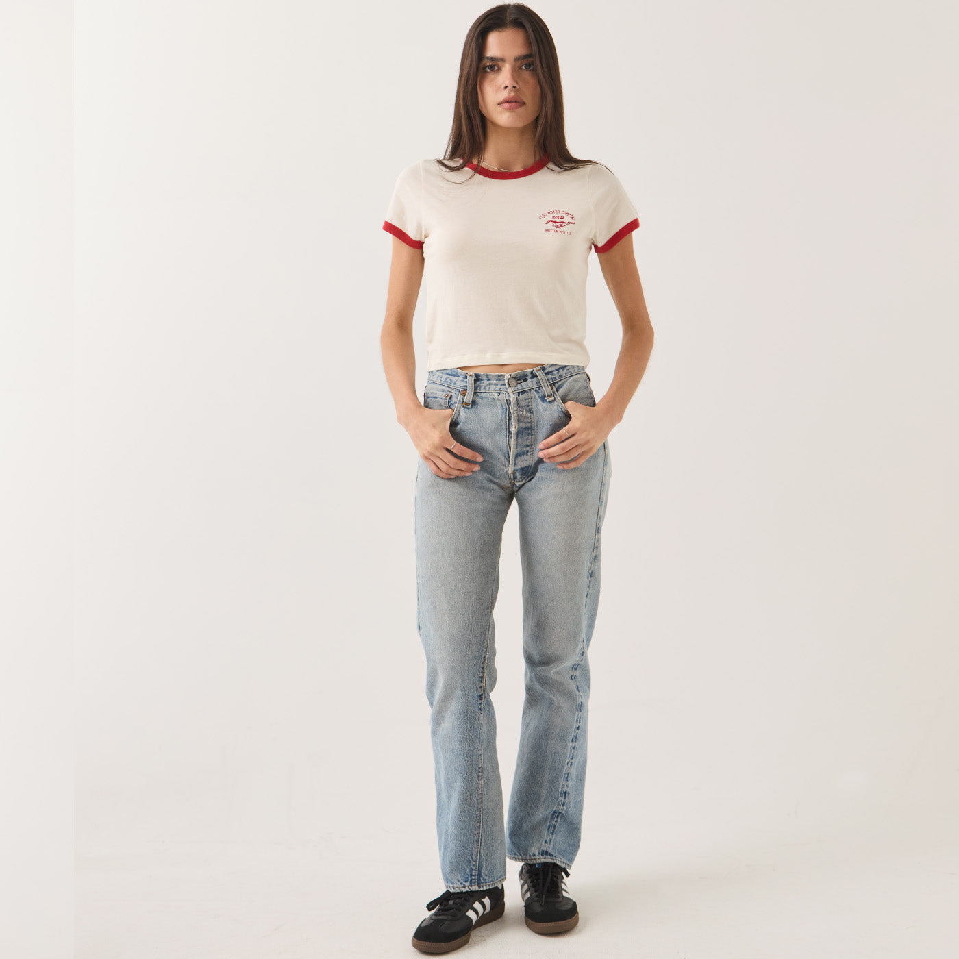 A woman modeling a cream ringer t-shirt with red trim and a small red graphic on the chest, paired with light-wash blue jeans and black sneakers, against a plain light gray studio background.