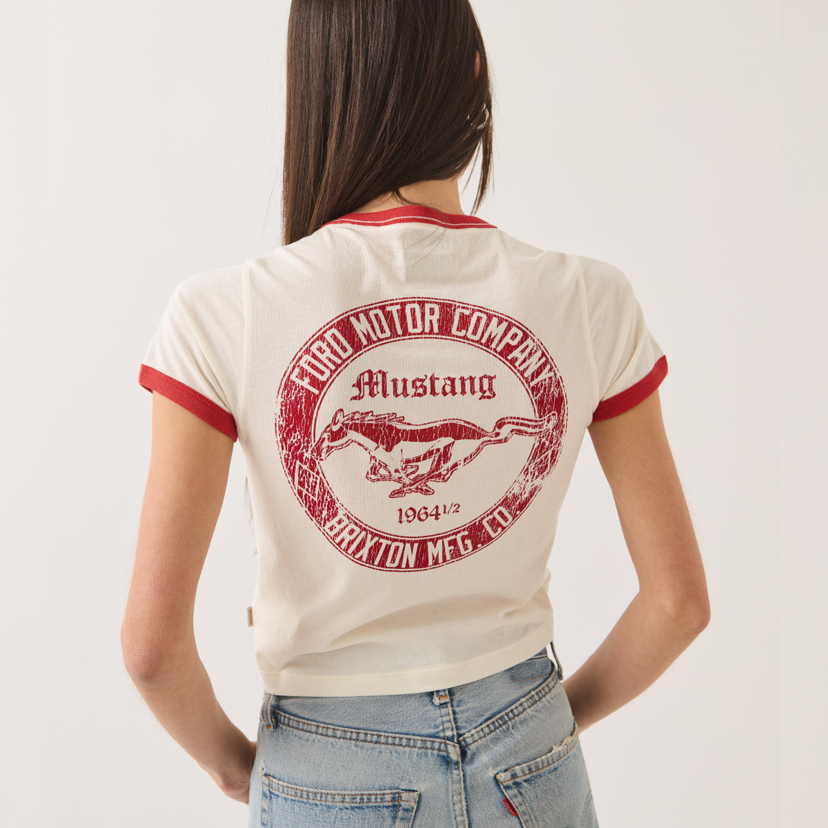 A woman shown from the back wearing a cream ringer t-shirt with red trim and a large red circular 'Ford Motor Company Mustang' graphic on the back, paired with light blue jeans against a plain white background.