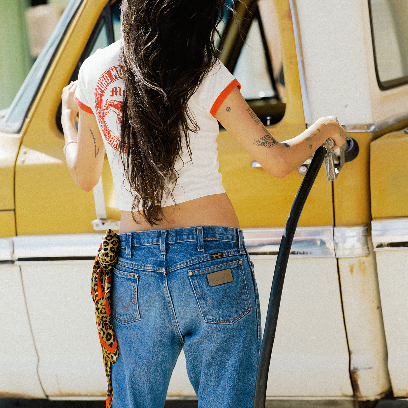 A woman viewed from behind wearing a white ringer t-shirt with orange trim and a red graphic, paired with blue jeans. She is standing next to a yellow and white vehicle, holding a black hose.