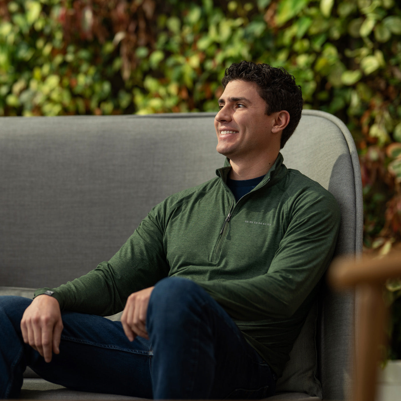 Man sitting on a gray couch wearing a green Bronco jacket with a blurred green plant background
