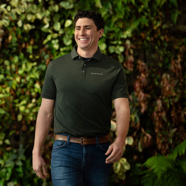 Man wearing a dark green Bronco polo shirt with text, standing in front of a leafy green background