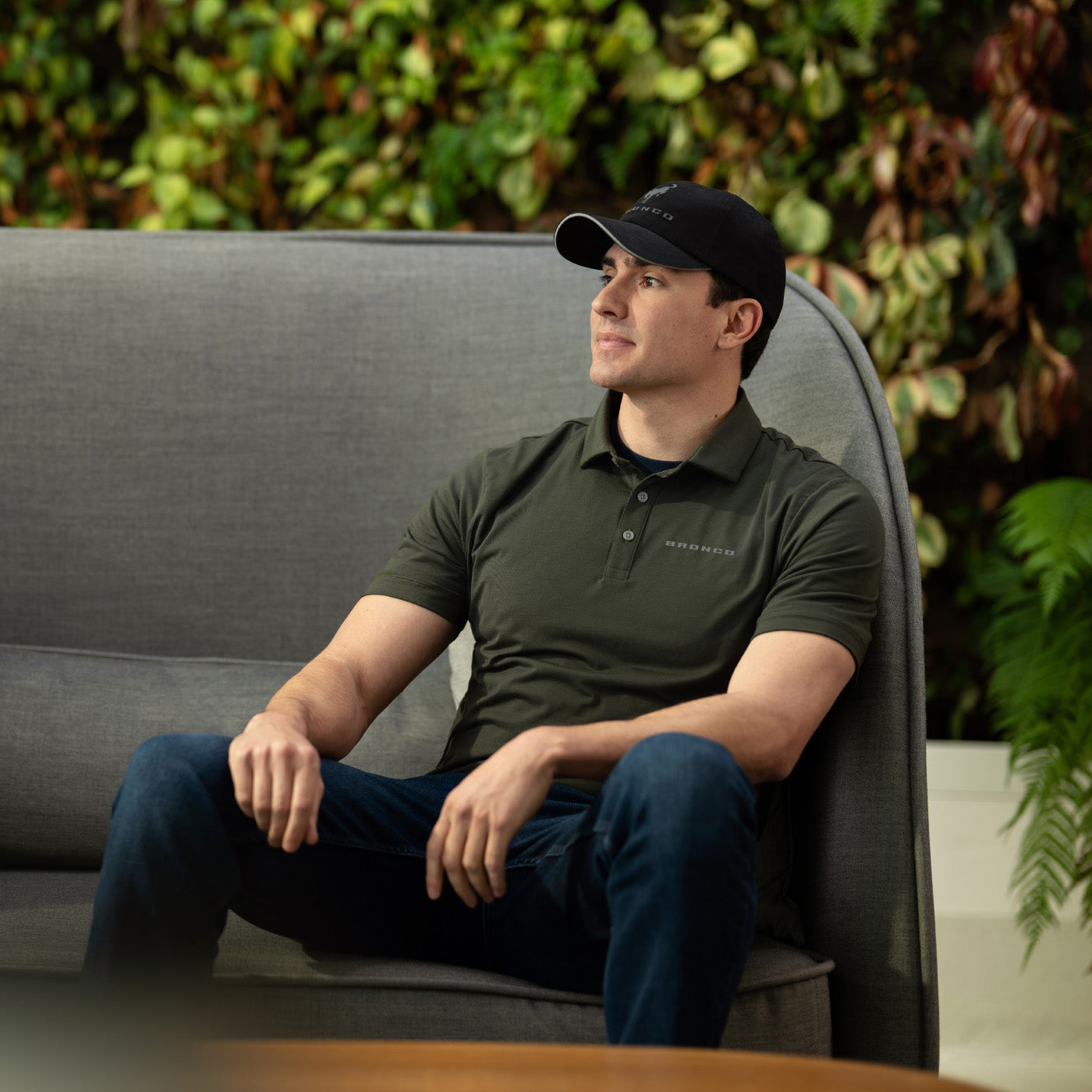 Man sitting on a gray couch wearing a green polo shirt and black cap, with a green plant wall in the background.