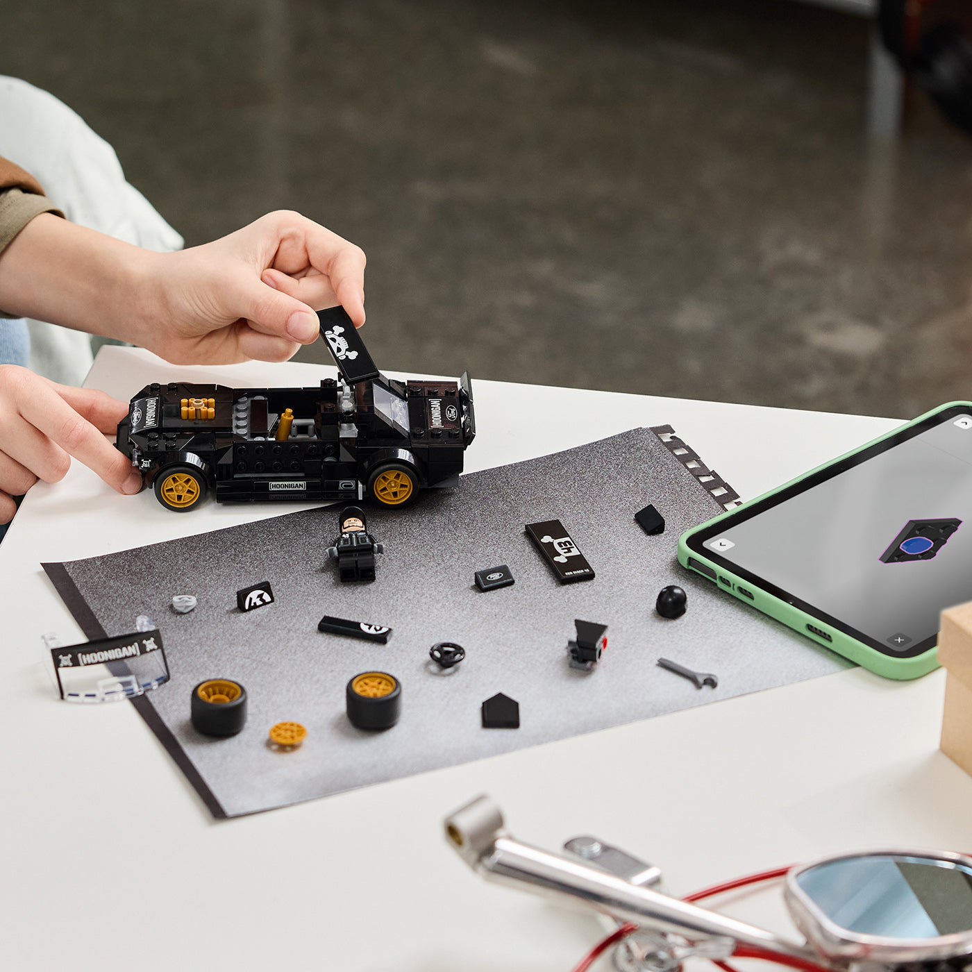 Person assembling a small vehicle model with scattered parts on a table, using a smartphone for reference.