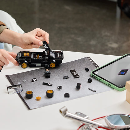 Person assembling a small vehicle model with scattered parts on a table, using a smartphone for reference.
