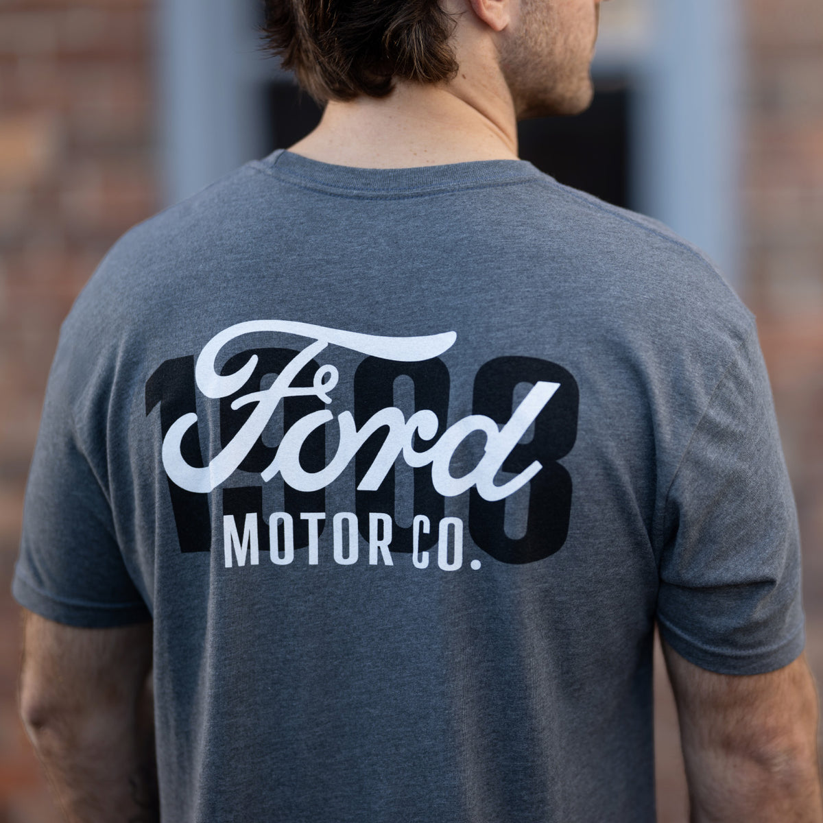 A man viewed from behind wearing a heather dark gray t-shirt with a large white and black Ford Motor Co. graphic on the back, set against a blurred brick wall background.
