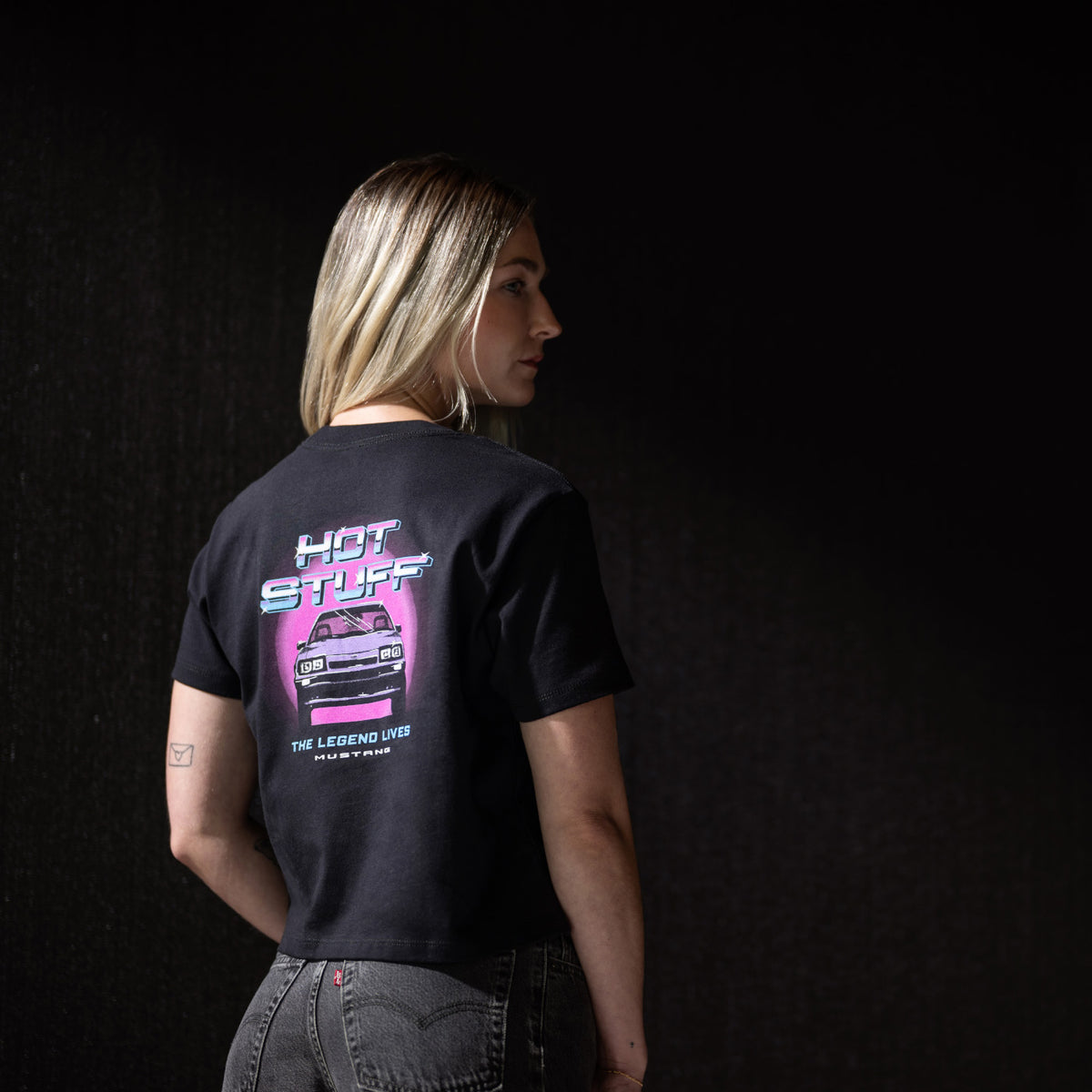 A woman shown from the back wearing a black graphic t-shirt with a pink and blue 'HOT STUFF' car design, paired with gray jeans, set against a dark studio background.