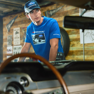 A man in a Ford Racing blue t-shirt and matching cap stands in a garage setting with a vintage car in the foreground.