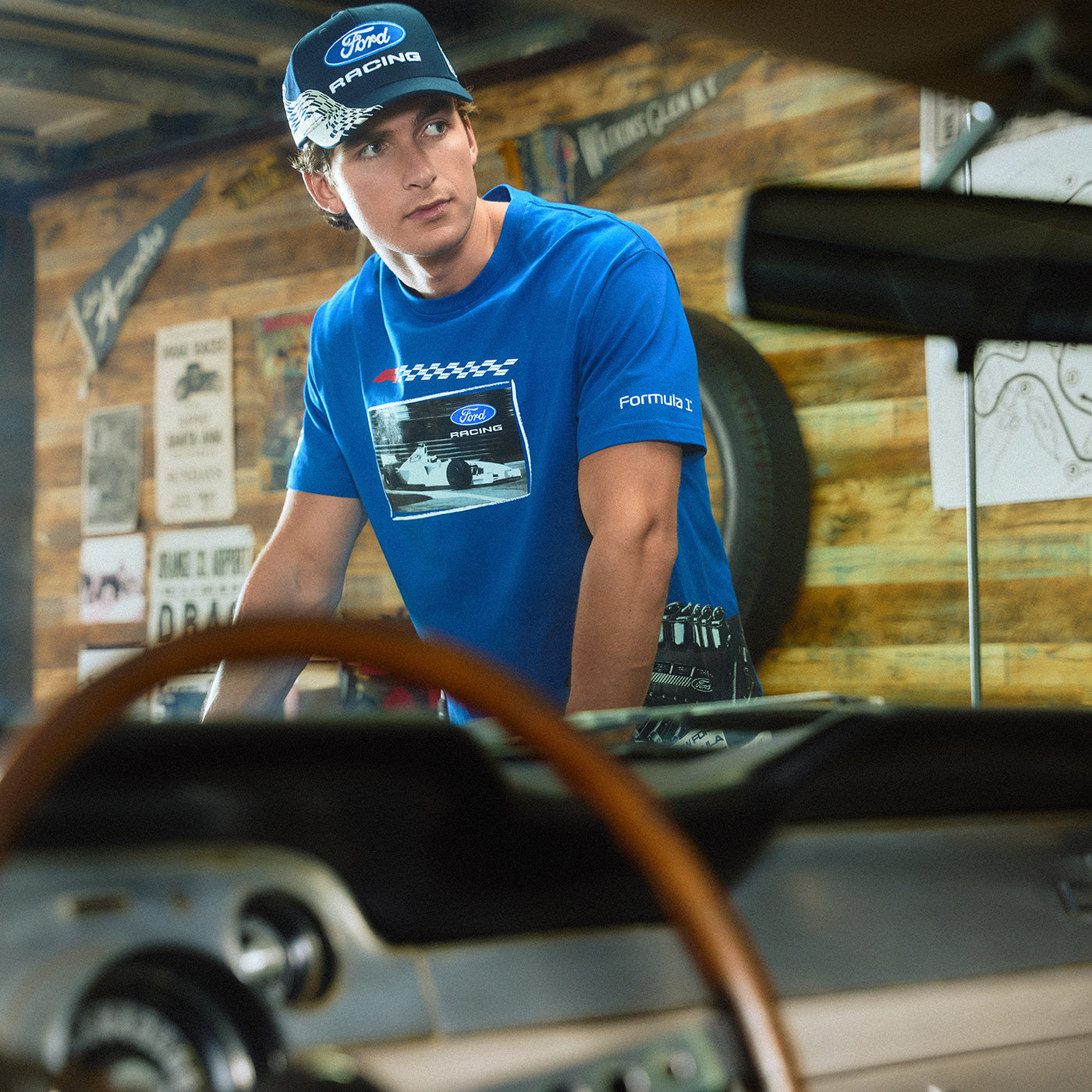 A man in a Ford Racing blue t-shirt and matching cap stands in a garage setting with a vintage car in the foreground.