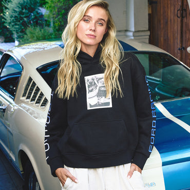 A blonde woman wearing a black pullover hoodie with a graphic print on the chest and 'FORD RACING' printed down the sleeve, standing in front of a blue and white classic Ford Mustang car.