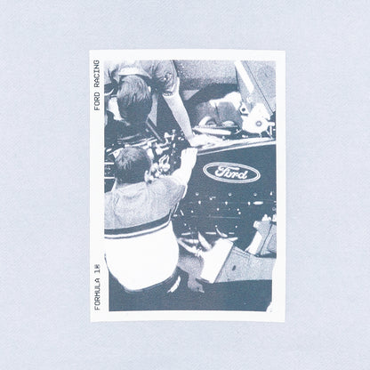 A vintage black and white Formula 1 Ford racing sticker or print, depicting drivers working on a car with the Ford logo visible, set against a light blue background.