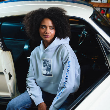 A woman with an afro sits in the open door of a white classic car, wearing a light blue hoodie with a black-and-white graphic on the chest and 'DEBATING' printed vertically in blue on the sleeve.