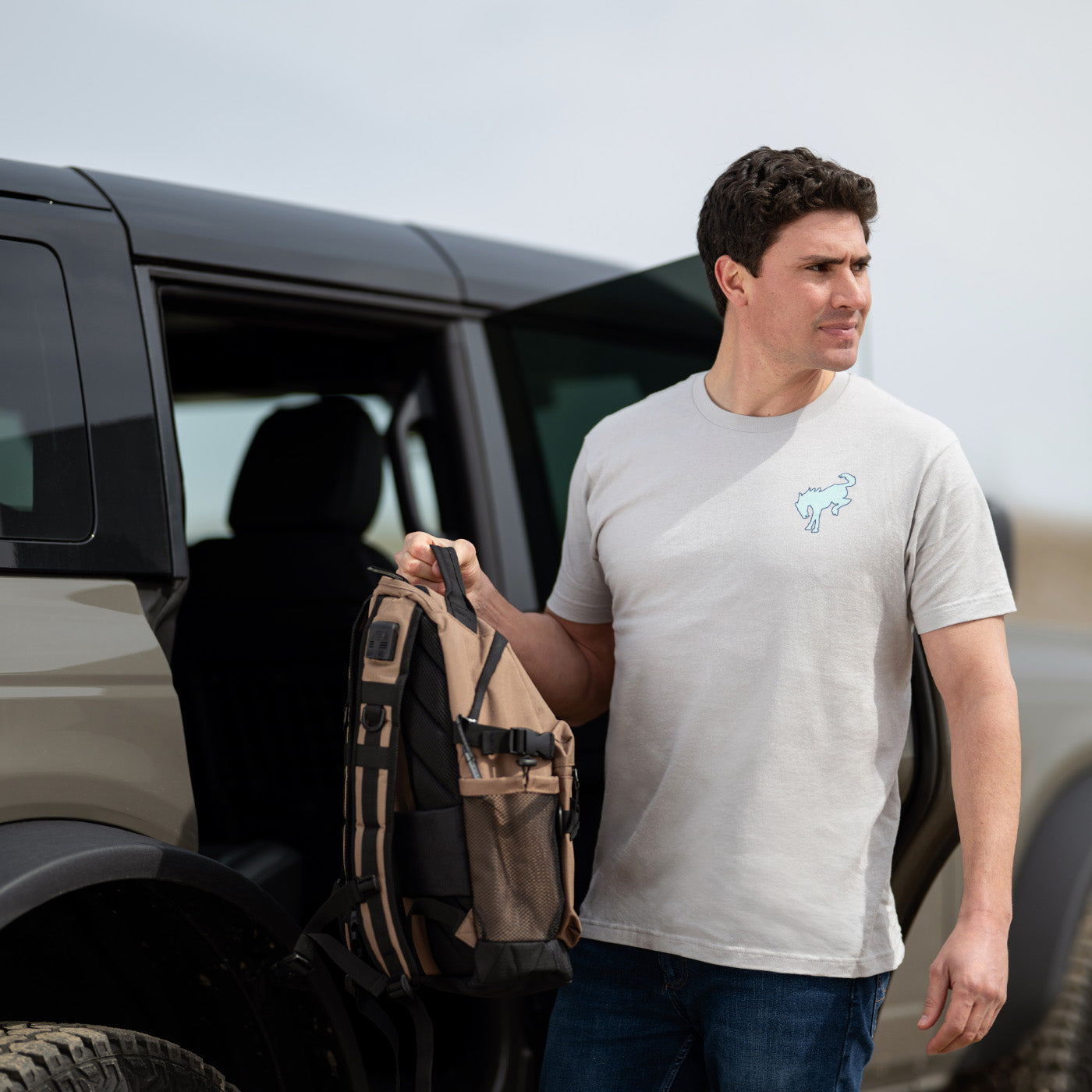 Man holding a bronco backpack next to a bronco vehicle in an outdoor setting