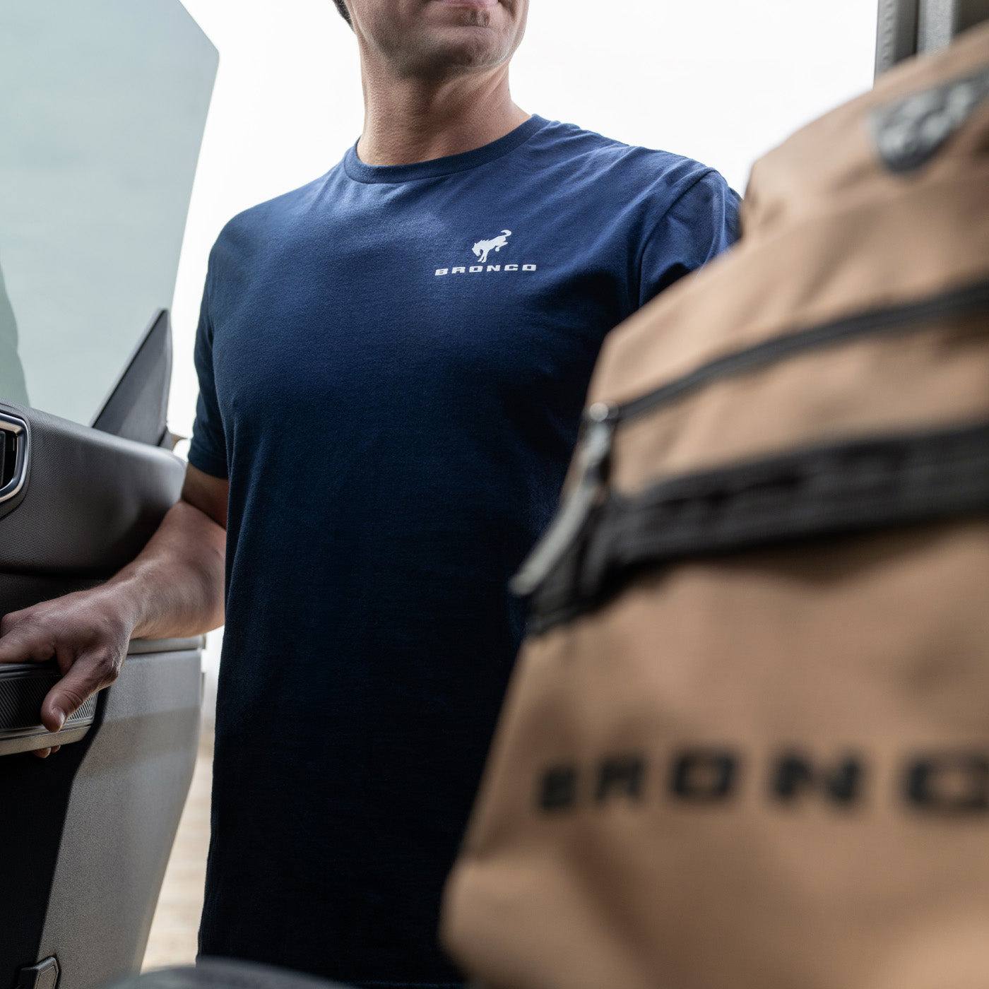 Person wearing a blue t-shirt with 'Bronco' logo opening a car door, with a brown bag featuring 'Bronco' branding in the foreground.