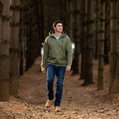 Man walking through a forest wearing a green jacket and blue jeans.