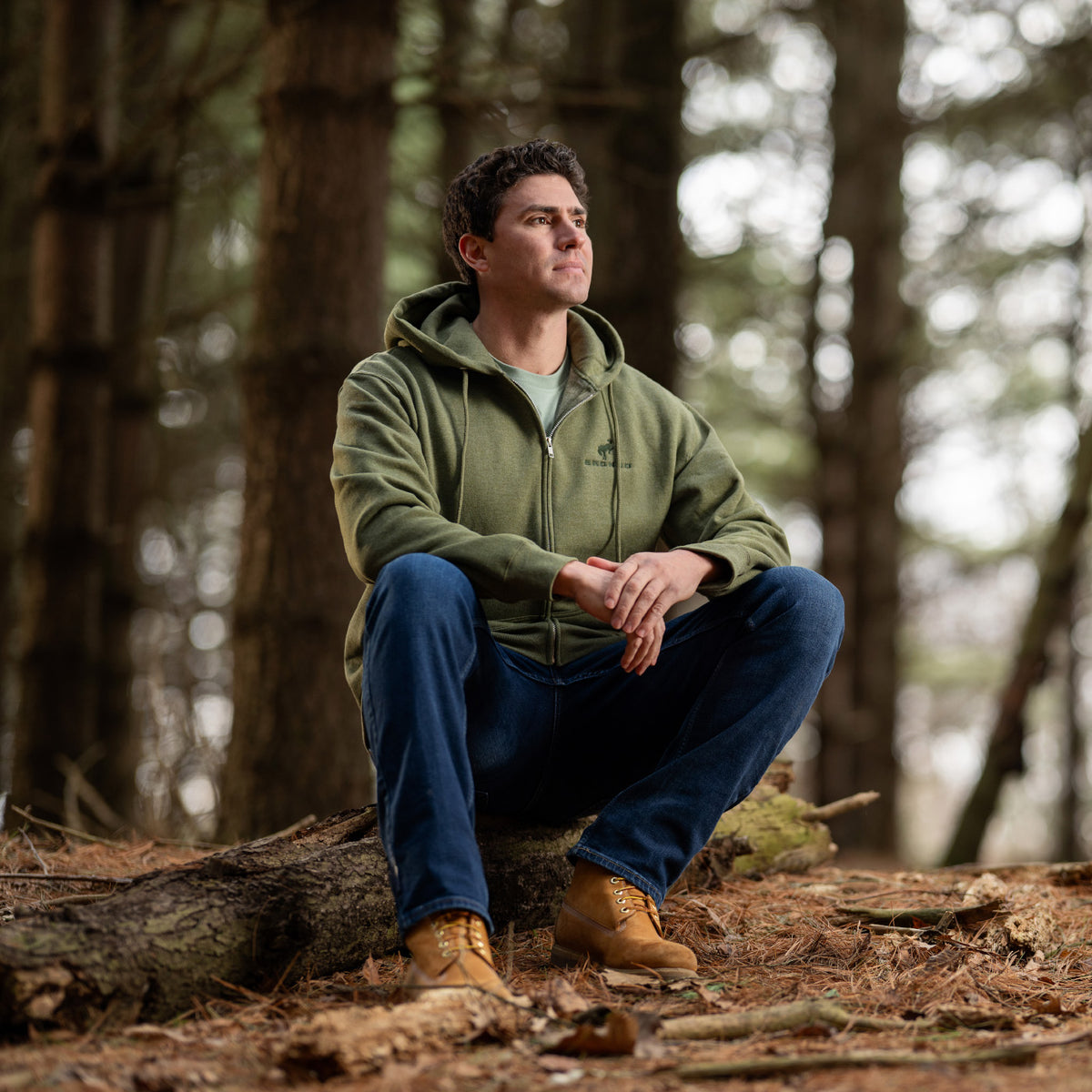 Man sitting on a log in a forest wearing a green hoodie and blue jeans.