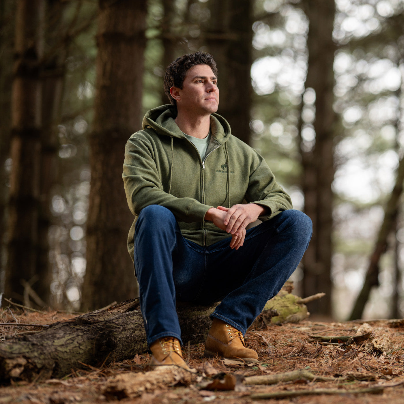 Man sitting on a log in a forest wearing a green hoodie and blue jeans.