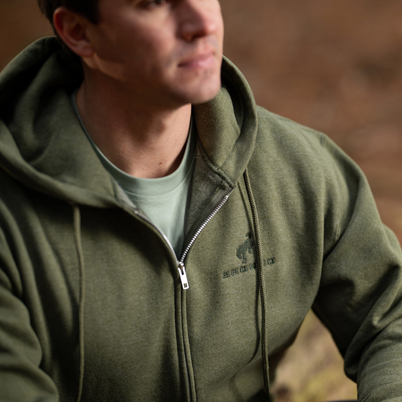 Person wearing a green hoodie with a logo, standing against a blurred natural background