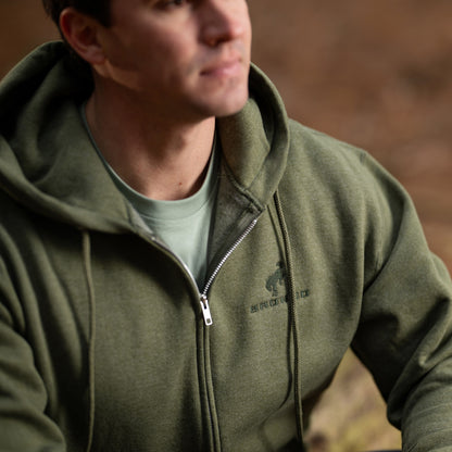 Person wearing a green hoodie with a logo, standing against a blurred natural background