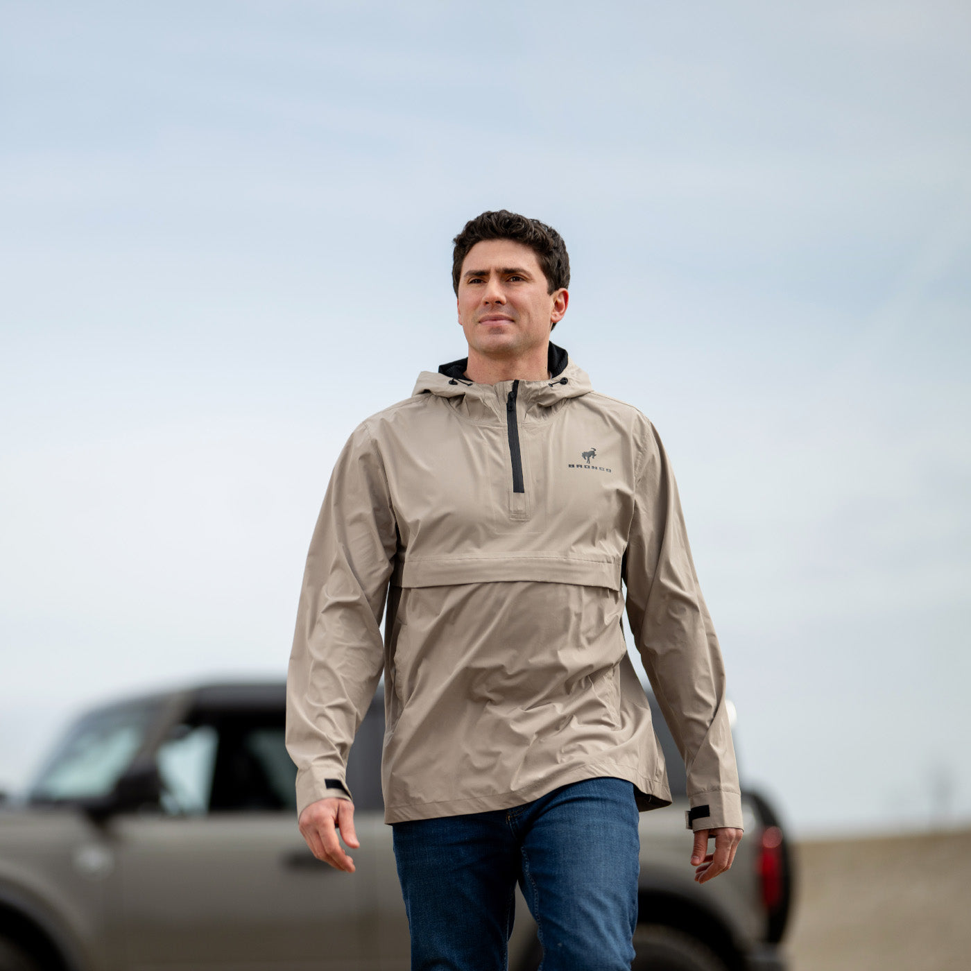 Man wearing a beige Bronco jacket with a logo, standing in front of a vehicle with a clear sky background