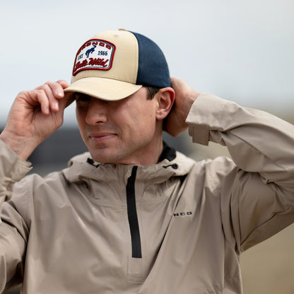 Man wearing a cap and beige jacket with a brand logo on a neutral background