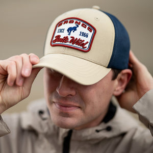 Man wearing a cap with a 'Bronco Since 1966' logo, holding it up to his head.