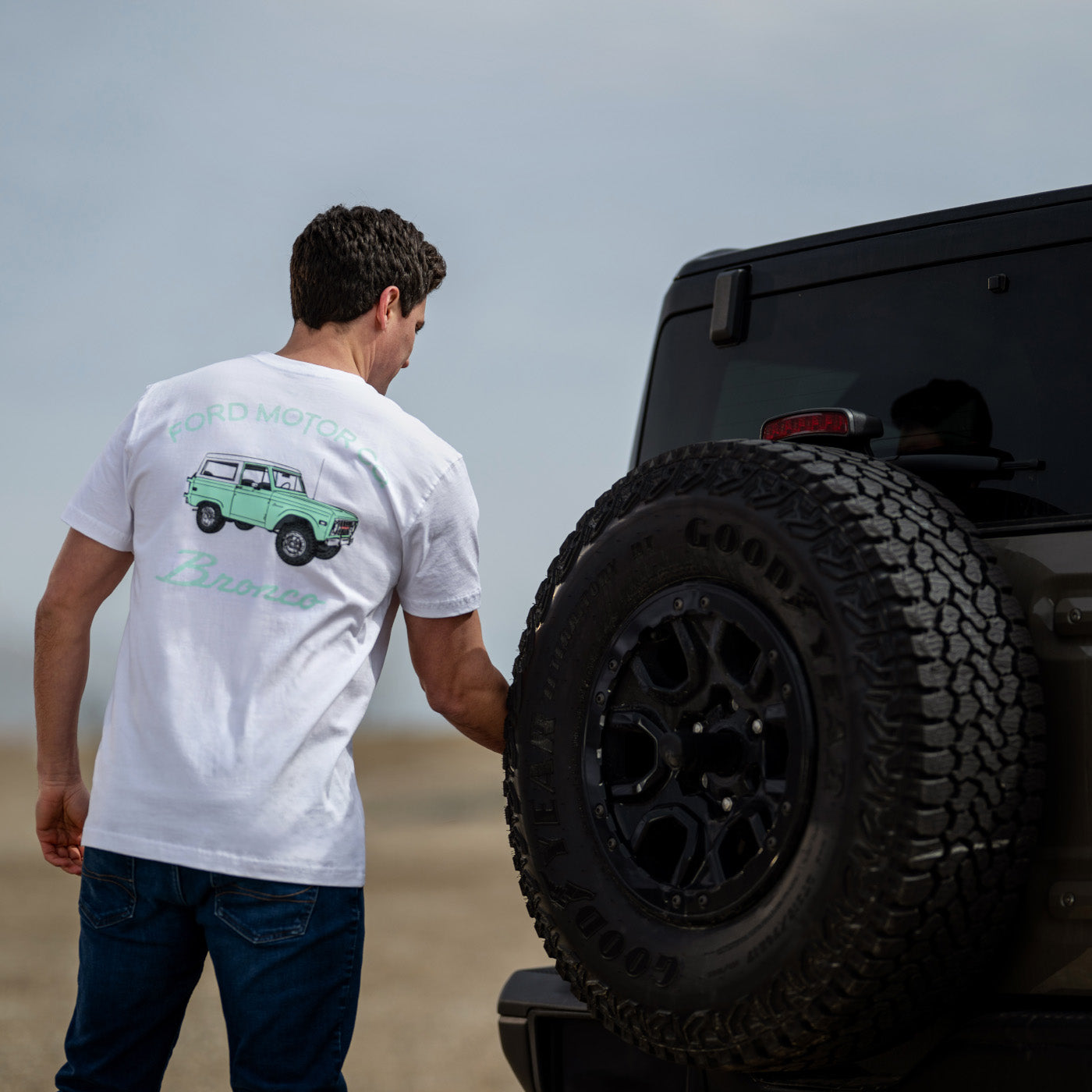 Man wearing a white t-shirt with a graphic design, standing next to a black off-road vehicle on a beach.