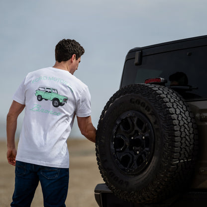 Man wearing a white t-shirt with a graphic design, standing next to a black off-road vehicle on a beach.