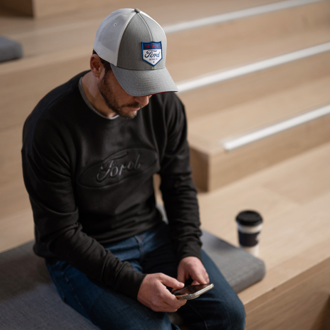 Man wearing a Ford cap and shirt sitting on steps using a phone with a coffee cup beside him.