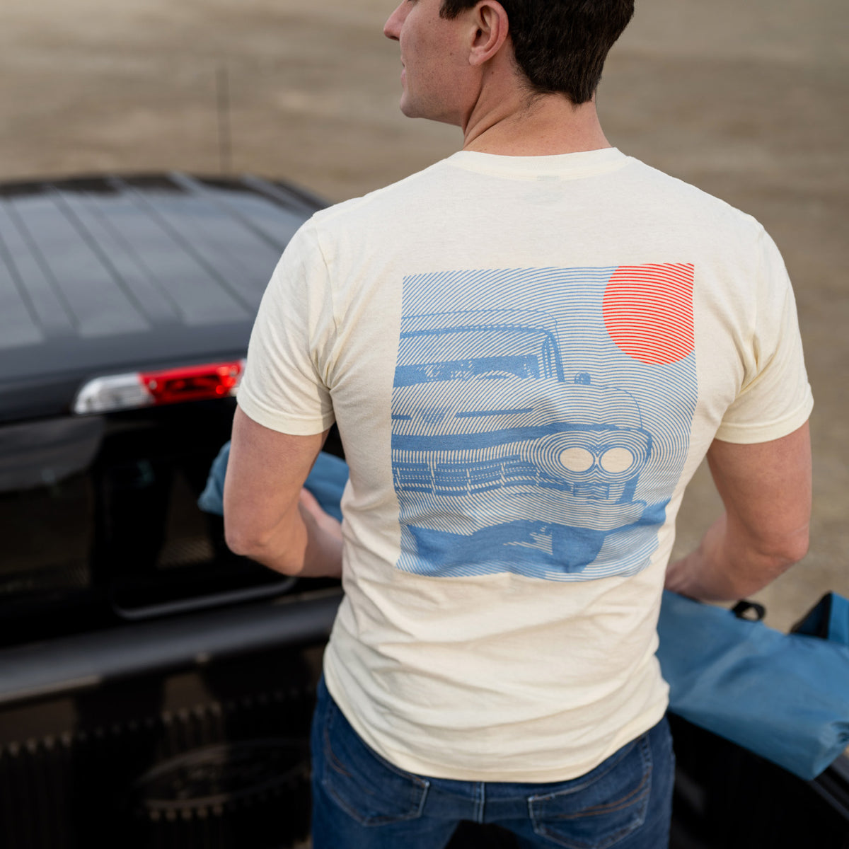 Man wearing a t-shirt with a graphic design of a car and sun, standing next to a car.