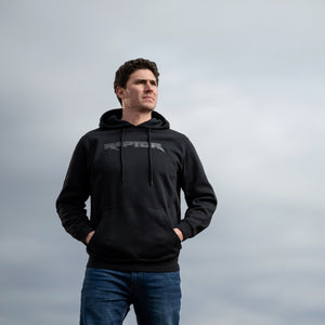 Man wearing a black raptor hoodie with a brand logo against a gray sky