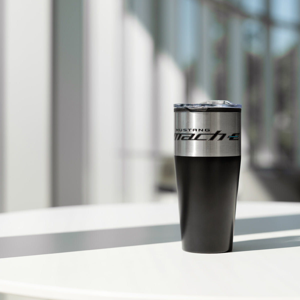 A black insulated travel tumbler with a silver metallic band reading 'HUSTLING MACH E' stands on a white table in a bright, modern indoor setting with blurred vertical architectural lines in the background.