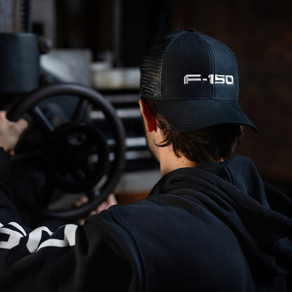 A man wearing a black trucker-style cap with 'F-150' embroidered in white on the front, viewed from behind as he sits in a vehicle interior near a steering wheel.