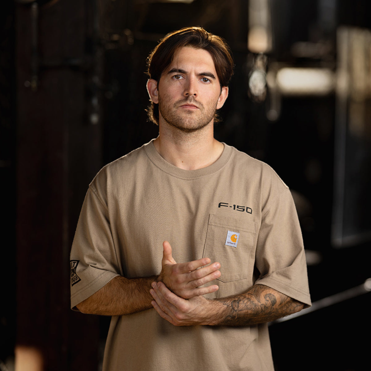 A man wearing a tan Carhartt F-150 short-sleeve work shirt stands in a dark, industrial setting with his hands clasped together. The shirt features the 'F-150' logo on the chest pocket and the signature Carhartt logo patch.