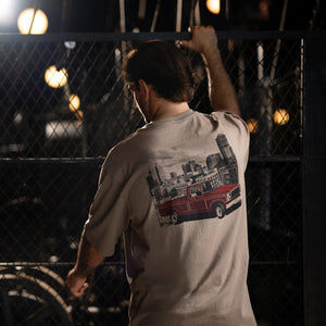 A man viewed from behind wearing a beige graphic t-shirt with a red vintage truck and city skyline print on the back, standing in front of a chain-link fence at night.