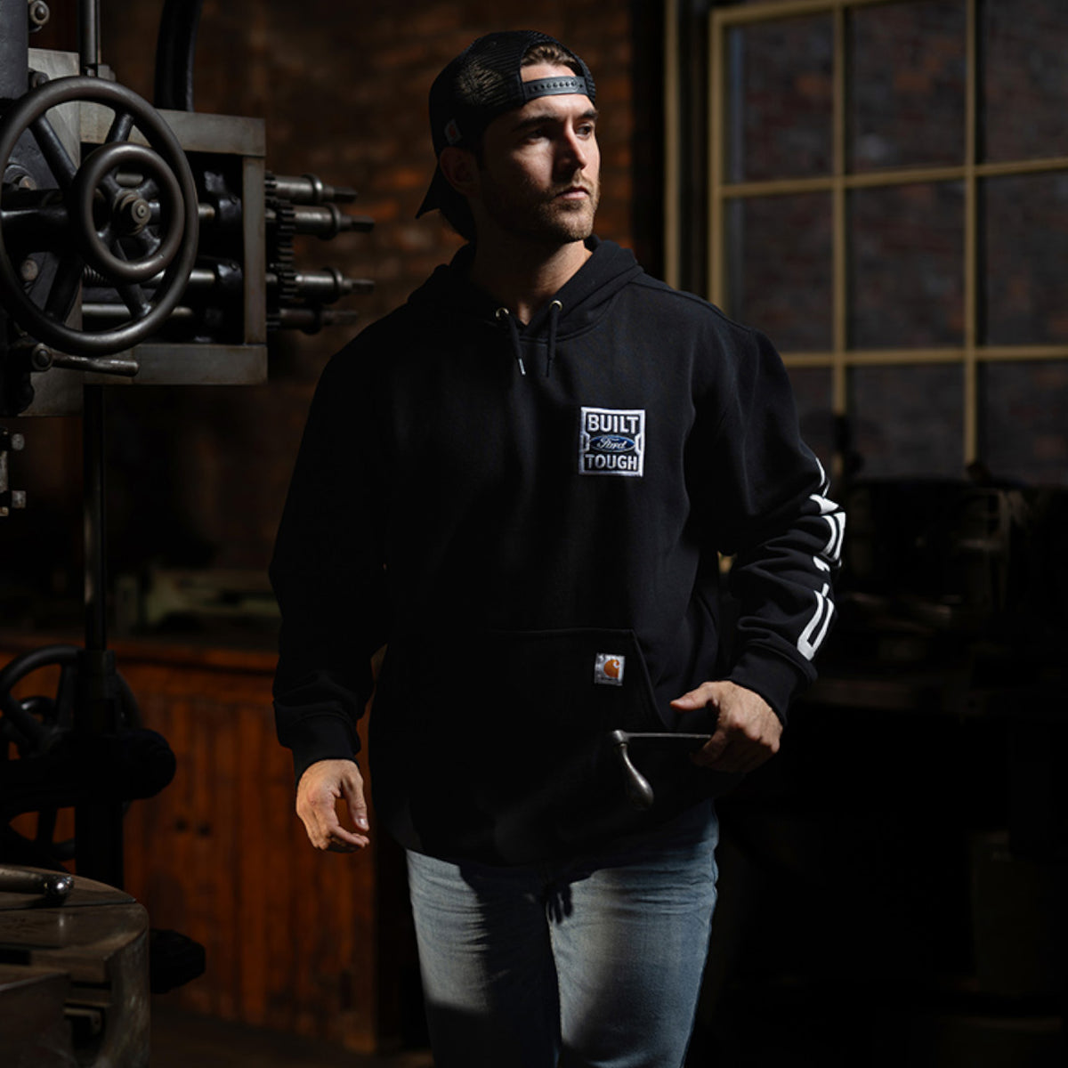 A man wearing a black Carhartt hoodie with a white 'Built Tough' logo on the chest, standing in a dimly lit workshop environment.