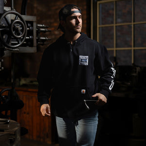 A man wearing a black Carhartt hoodie with a white 'Built Tough' logo on the chest, standing in a dimly lit workshop environment.