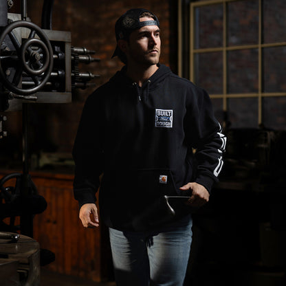 A man wearing a black Carhartt hoodie with a white 'Built Tough' logo on the chest, standing in a dimly lit workshop environment.
