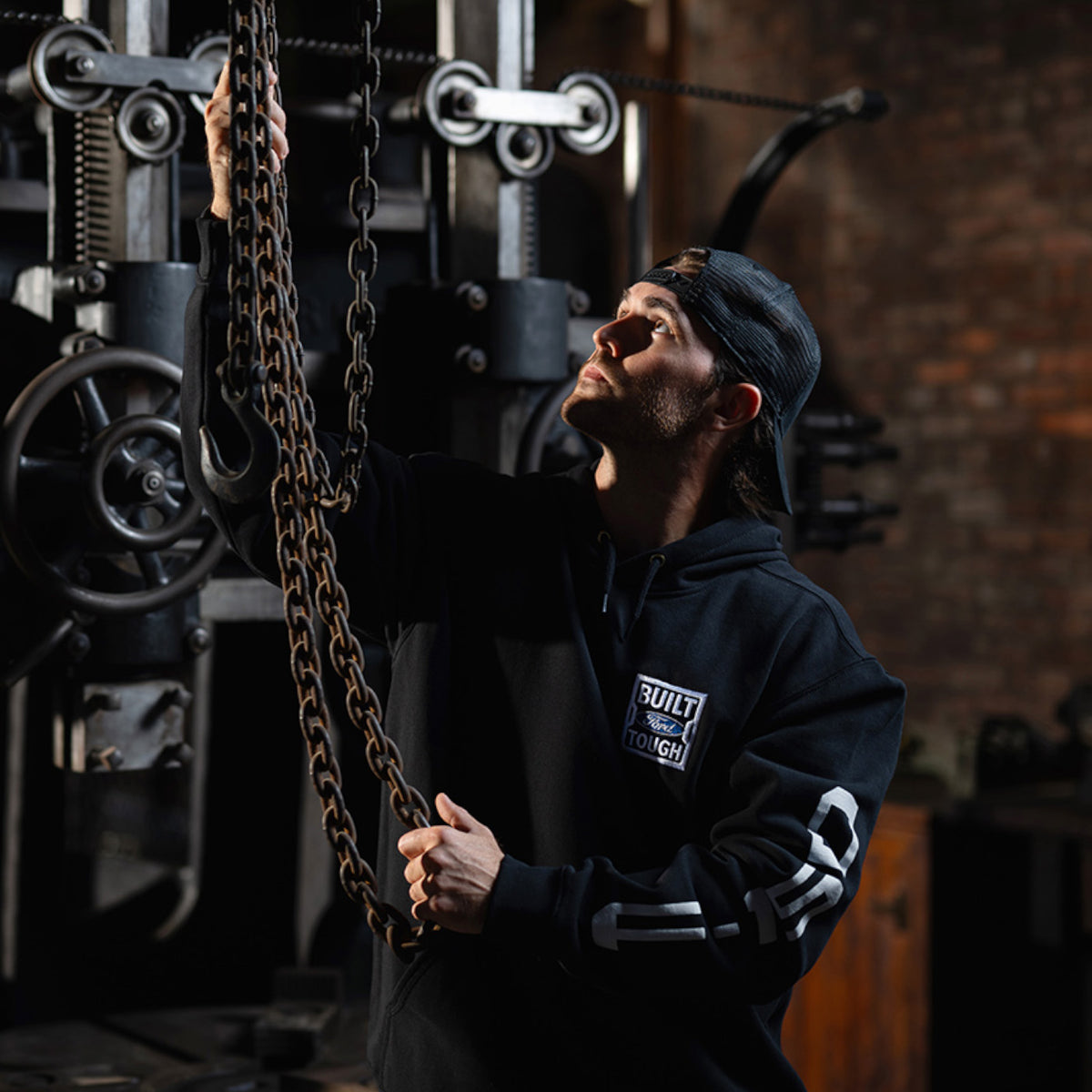 A man in a gym wearing a black hoodie with a 'Built Tough' logo, looking up at a hanging chain attached to a weight machine.